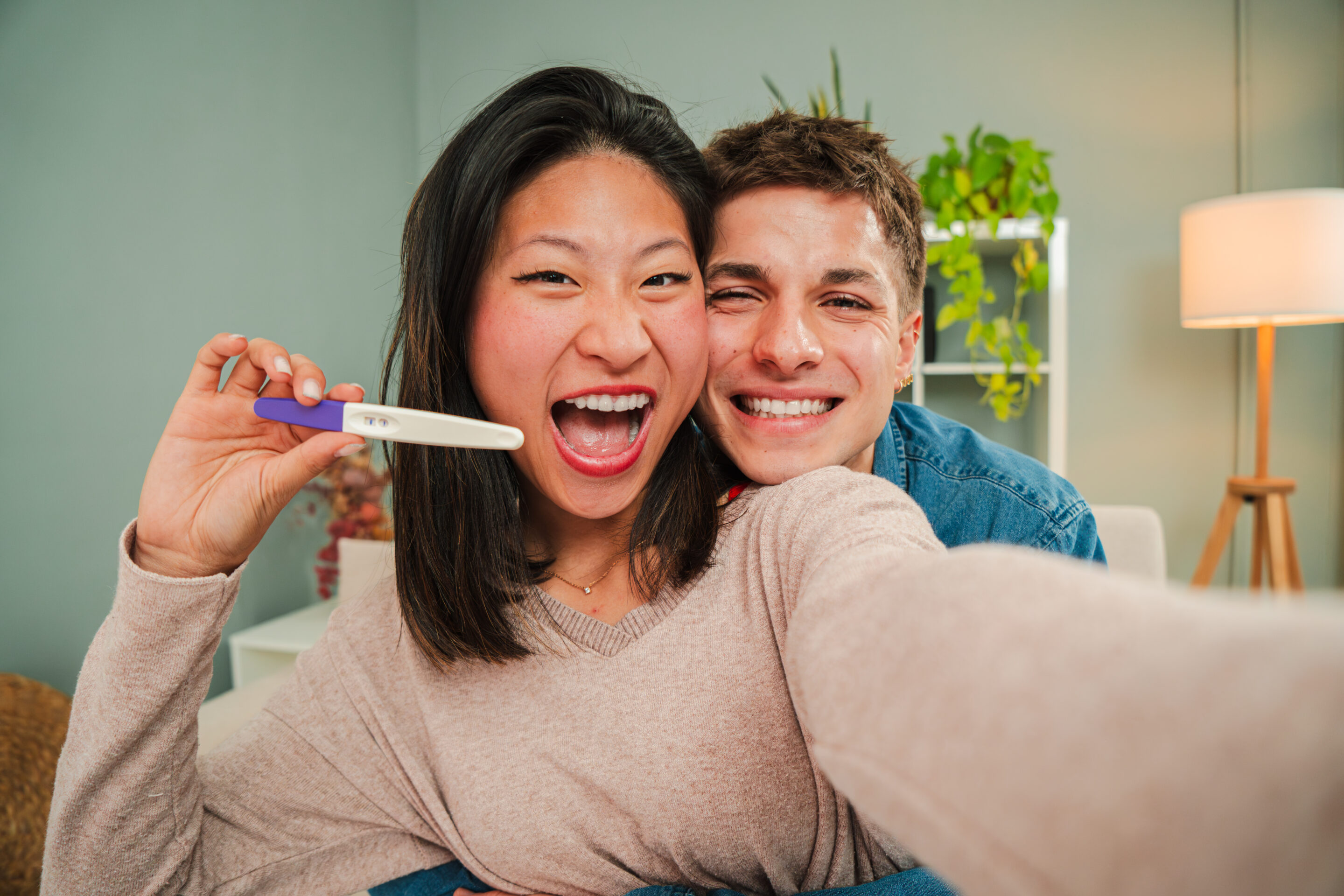 A young couple hugging and showing a positive pregnancy test to the camera at home. Mother and father feel happy about their motherhood and forming a family. Two joyful people expecting a first baby